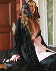 A model sitting on a sofa wearing black coat, pink shirt