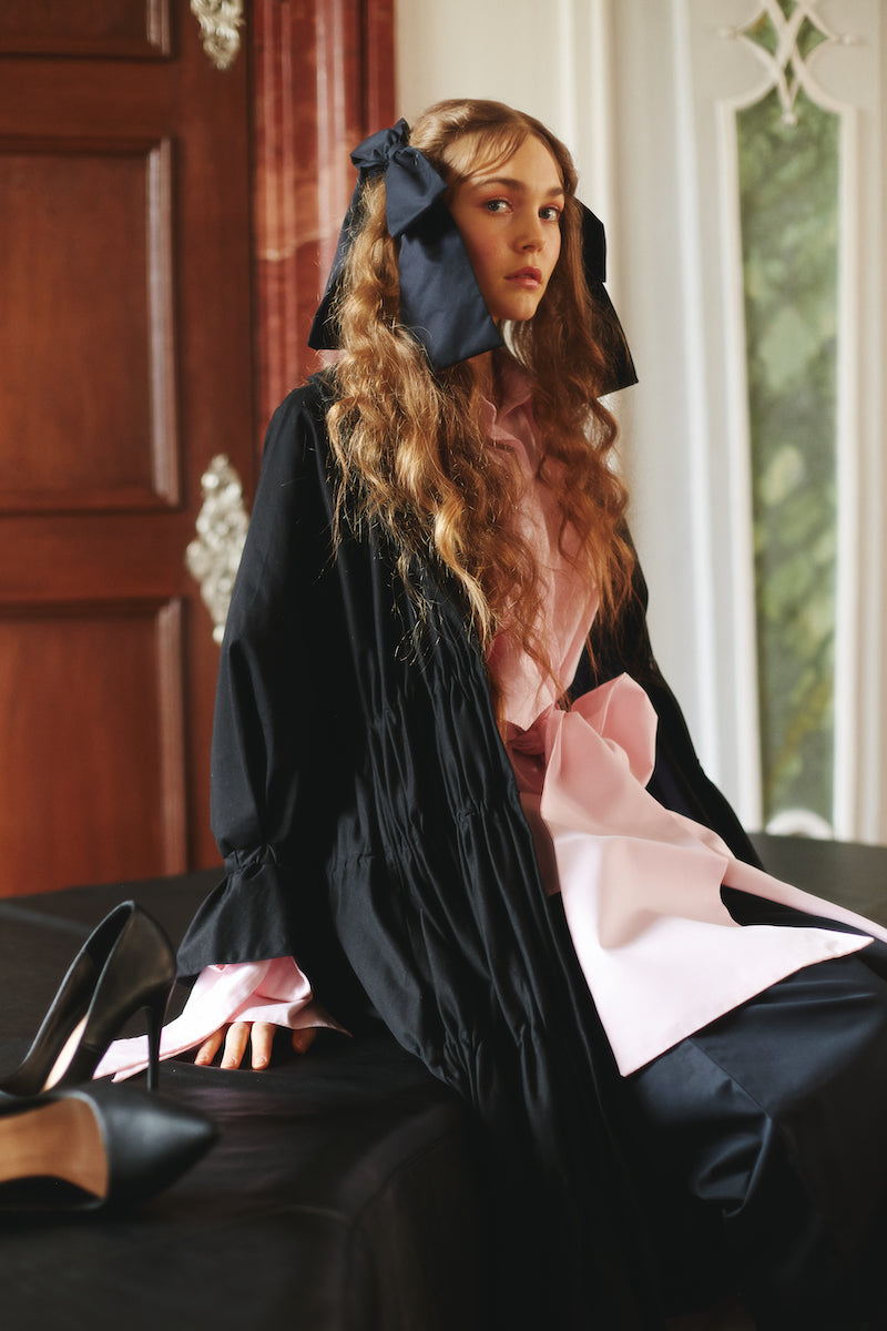 A model sitting on a sofa wearing black coat, pink shirt