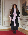 A model is wearing a black coat, pink shirt whit a huge ribbon and japanese like trousers. She has also 2 black ribbons in her hair and high heels.